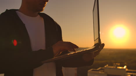 Close-up:-a-programmer's-hand-typing-on-a-laptop-keyboard-at-sunset-overlooking-the-roof.-A-businessman-works-remotely.-Freelancer-performs-work-on-vacation